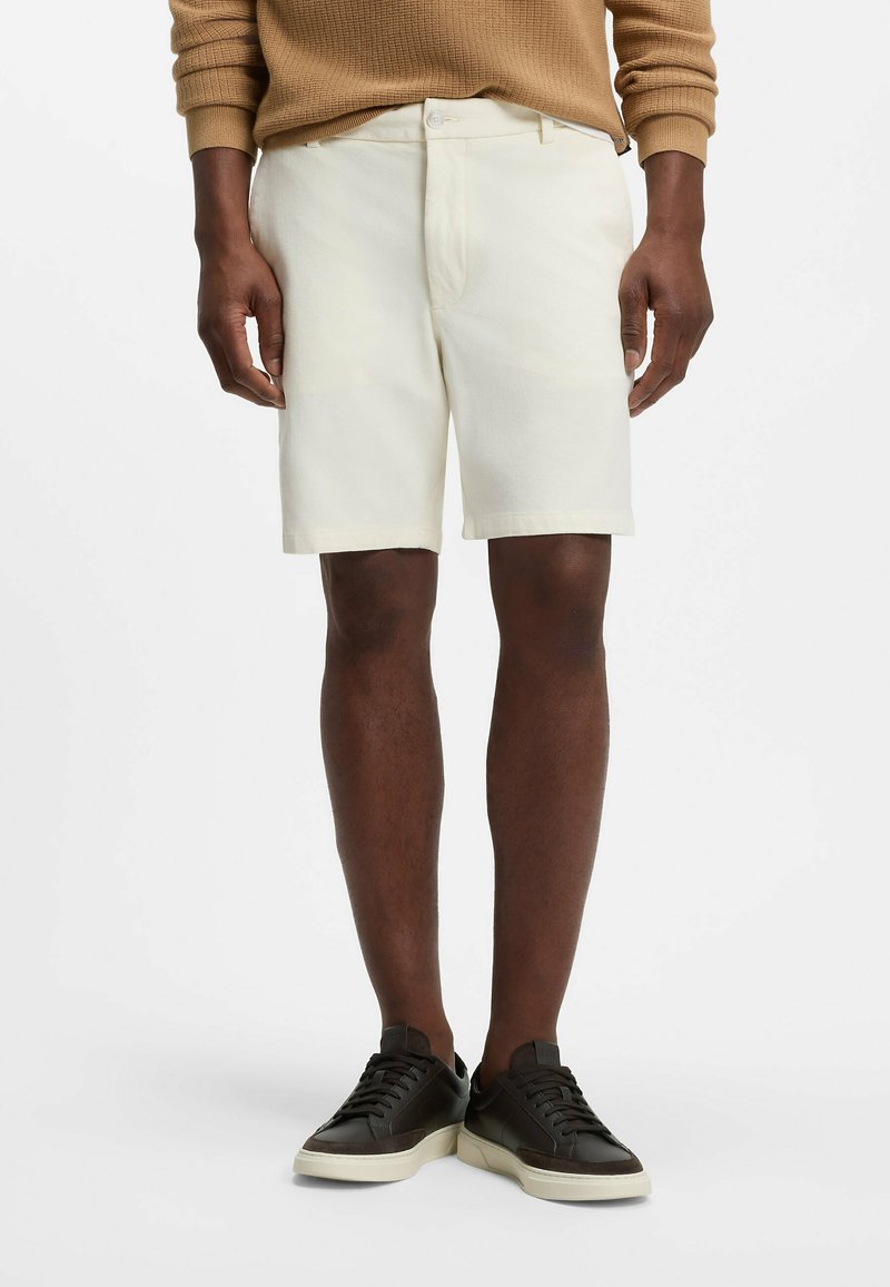 Homme portant un short beige, un pull marron à manches longues et des baskets noires avec semelles blanches, debout devant un fond blanc.