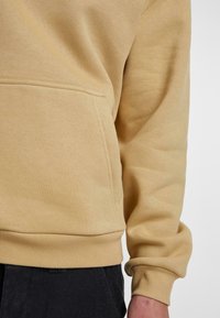 Sweat-shirt beige avec ourlet et poignets côtelés porté par une personne, montrant une poche avant et une partie d'un pantalon noir en dessous.