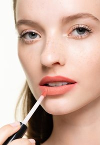 Matt flytande läppstift appliceras med en doe-foot applikator; mjuk persikofärg, slät textur, accentuerad av lätta fräknar på huden.