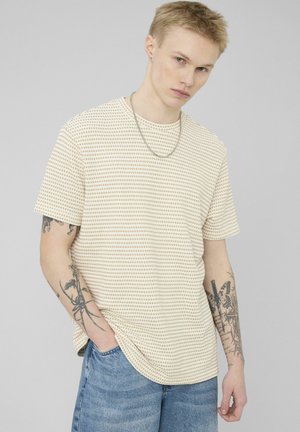 Jeune homme aux cheveux blonds portant une chemise beige à motifs à manches courtes, un collier en chaîne, un jean bleu et des tatouages visibles sur les bras.