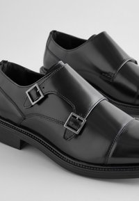 Chaussures en cuir noir à finition lisse. Dotées de deux brides avec boucles et d'un design structuré. Comprend un petit talon empilé et des coutures décoratives.