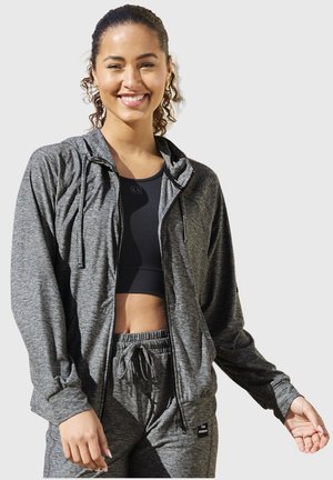 Smiling woman wearing gray zip-up hoodie, matching sweatpants, and black sports bra against a plain light background.