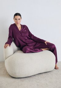 Lila samettinen pyjamapaketti, jossa on teksturoitu kuvio, täydentää kodikas, vaalean kreemi värinen bouclé-tyyppinen nojatuoli, jossa on pyöreä muoto.