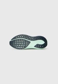 Semelle de chaussure de course avec un motif en caoutchouc noir pour l'adhérence sur un fond vert clair, vue de dessous sur un fond uni.