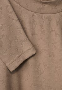 Beige Stoff mit einer weichen Textur zeigt eingeprägte Muster von stilisierten Tieren. Der runde Ausschnitt und die umgeschlagenen Ärmel sind sichtbar.
