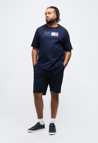 Άνδρας με dreadlocks που φοράει μπλε μπλουζάκι Tommy Hilfiger, μπλε σορτς, άσπρες κάλτσες και μπλε αθλητικά παπούτσια, στέκεται με τα χέρια στις τσέπες.