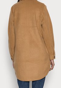 Lang brun frakke i uldblanding med afrundet kant, knappede manchetter og klassisk krave, der har en glat tekstur og løs pasform.