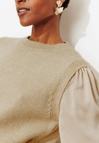 Pull beige en maille avec accents scintillants, doté d'un col côtelé et d'épaules texturées, accompagné d'un chemisier crème à manches bouffantes.
