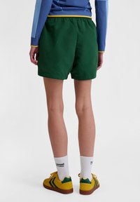 Gröna shorts med elastisk midja, med en slät yta och snitt ovanför knäna, kombinerade med gula skor och vita strumpor.