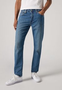 Modré džínové kalhoty s rovnými nohavicemi, s pětiprvkovým designem a zapínáním na knoflíky; doplněné bílými teniskami na neutrálním pozadí.