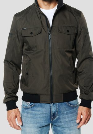 Mann trägt eine dunkelgrüne Jacke mit Reißverschluss und knöpfbaren Brusttaschen sowie blaue Jeans, steht vor einem schlichten hellen Hintergrund.