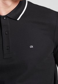 Svart pikétröja i texturerad bomull, med en delad vit krage, treknapps-knäppning och en liten vit "ck"-logotyp på bröstet.