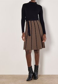 Femme portant un haut noir à manches longues côtelé, une jupe mi-longue à motifs avec des rayures noires verticales, et des bottines noires, debout à l'intérieur.