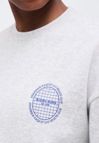 Κοντινή λήψη ενός ανοιχτόγκρι φούτερ με μπλε κυκλικό έμβλημα που δείχνει το πλέγμα ρακέτας τένις και το κείμενο «Stockholm Racquet and Health Club» και «Björn Borg Est. 1956.»