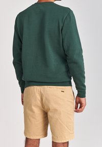 Felpa verde scuro con collo a giro, maniche lunghe e polsini a coste, abbinata a pantaloni shorts color khaki, che mostrano una vestibilità rilassata e una trama di cotone.