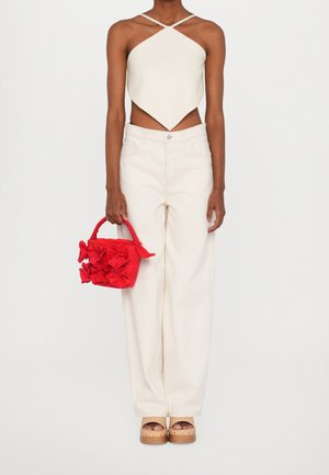 Femme portant un haut à col licou blanc et un pantalon à jambes larges, tenant un sac à main rouge texturé avec de grands nœuds, et des sandales à plateforme beiges.