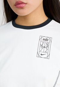 White t-shirt with black ribbed crew neck, featuring a front graphic with "nike", "1972", and laurel design. Soft fabric, casual fit.
