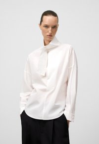 Camicia bianca in cotone con colletto alto, maniche lunghe e dettaglio a cravatta al collo. Presenta una vestibilità comoda e una texture liscia.