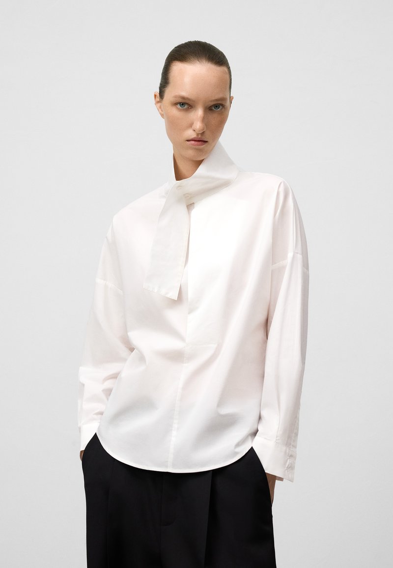 Chemise en coton blanche avec un col haut, des manches longues et un détail de nœud papillon au cou. Présente une coupe décontractée et une texture lisse.
