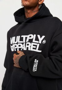 Felpa nera in cotone con tasca a marsupio. "MULTIPLY APPAREL" scritto in grandi lettere bianche sul davanti. Testo sulla manica con la scritta "IN FACTS WE TRUST."