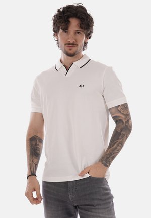 Hombre con el pelo rizado que lleva una camisa polo blanca con ribete negro y tatuajes en ambos brazos, de pie con una mano en el bolsillo.