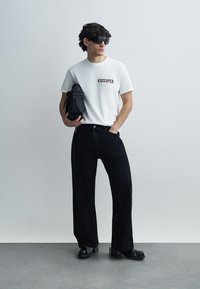 Λευκό μπλουζάκι με στάμπα "KIDSUPER", μαύρο παντελόνι τζιν καλοραμμένο και μαύρα παπούτσια με ασημένιες λεπτομέρειες. Κρατάει μια μαύρη τσάντα, στέκεται μπροστά σε έναν φωτεινό τοίχο.