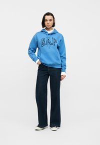 Jeune femme debout portant un sweat à capuche bleu de chez GAP, un jean large sombre, des baskets blanches, et les poignets d'une chemise blanche visibles sous le sweat.