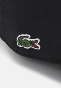 Tissu noir avec un logo de crocodile brodé en vert et rouge, entouré d'une bordure blanche, présentant une texture lisse et mate.