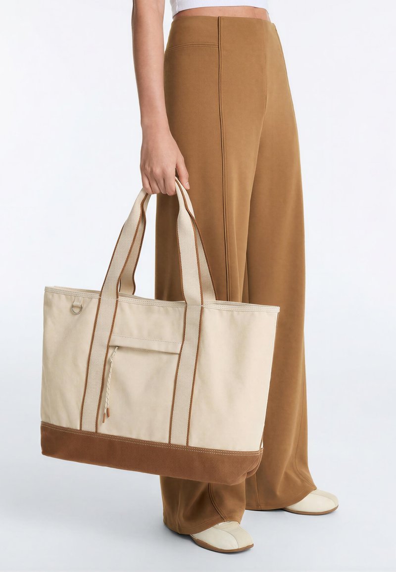 Personne tenant un grand sac cabas en toile beige avec des accents marron, portant un pantalon marron taille haute à jambes larges et des chaussures crème.