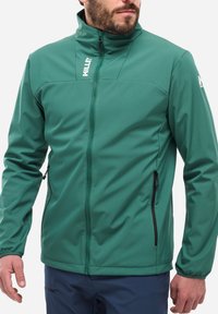 Veste softshell verte avec un col haut, une fermeture �éclair sur le devant et des poches latérales. Prend en charge un tissu lisse et texturé avec des fermetures éclair noires contrastantes.