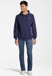 Sweatshirt blu navy con cappuccio e cordino, caratterizzato da un logo sul petto. Abbinato a jeans azzurri e sneakers bianche. Texture liscia.