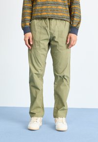 Light green cargo pants with elastic waistband, two side pockets, and a relaxed fit, paired with white sneakers on a blue background.