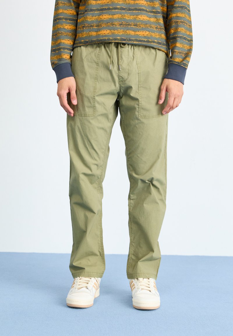 Light green cargo pants with elastic waistband, two side pockets, and a relaxed fit, paired with white sneakers on a blue background.