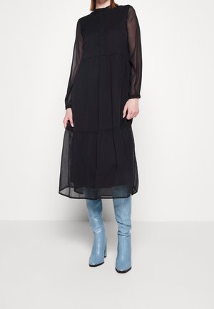 Personne portant une robe noire mi-longue avec des manches longues transparentes et des bottes hautes bleu ciel à talons, debout devant un fond blanc.