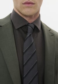 Blazer olive ajusté sur une chemise noire à chevrons sombres avec une cravate rayée charbon, mettant en valeur des textures et motifs distincts.