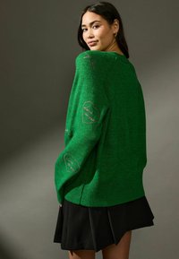 Maglione verde lavorato a maglia con maniche lunghe, orlo a coste e ricamo a forma di cuore sulle maniche. Modello che indossa una gonna nera.