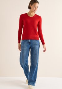 Roter, langärmeliger V-Ausschnitt-Pullover aus weichem Strickstoff, kombiniert mit hellblauen, geraden Jeans und beigen Sneakers.