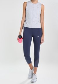 Top gris sin mangas, leggings azul marino con detalles de malla y zapatillas deportivas grises. Sosteniendo una botella de agua rosa.