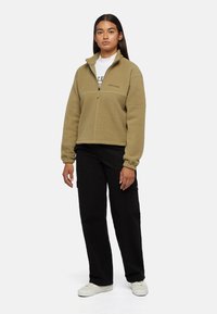 Sudadera de forro polar en verde olivo, con medio cierre, puños elásticos y un logo en el pecho. Combinada con pantalones cargo negros y zapatillas blancas.