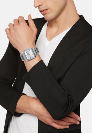Montre rectangulaire argentée avec un cadran blanc, bracelet en acier inoxydable et marqueurs d'heure minimalistes. Portée au poignet, associée à un blazer noir.