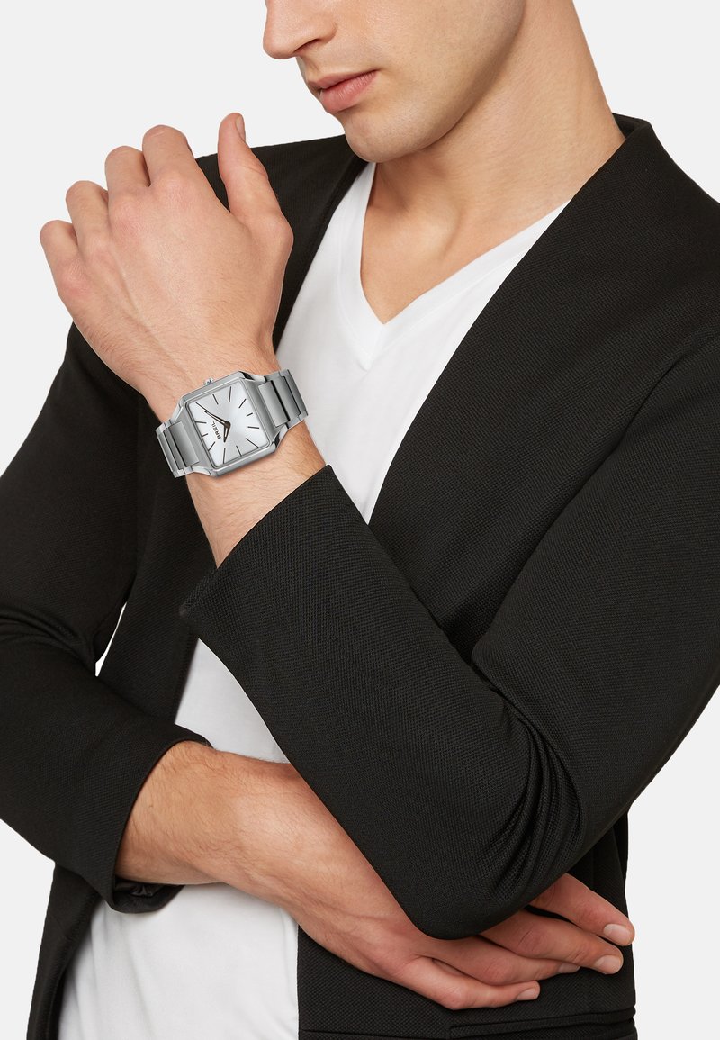 Sølv rektangulært ur med en hvid urskive, rustfrit stålarmbånd og minimaliske timemarkeringer. Båret på et håndled, parret med en sort blazer.