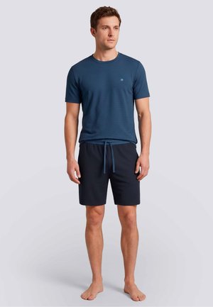 Uomo in piedi scalzo con una maglietta blu a maniche corte e pantaloncini blu scuro con coulisse su uno sfondo uniforme.