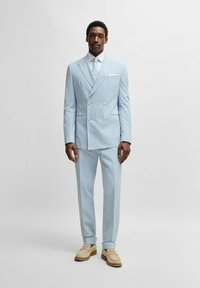Ljusblå dubbelknäppt kostym med texturerad tyg, matchande slips, vit skjorta och beige loafers. Kostymen har två framfickor.