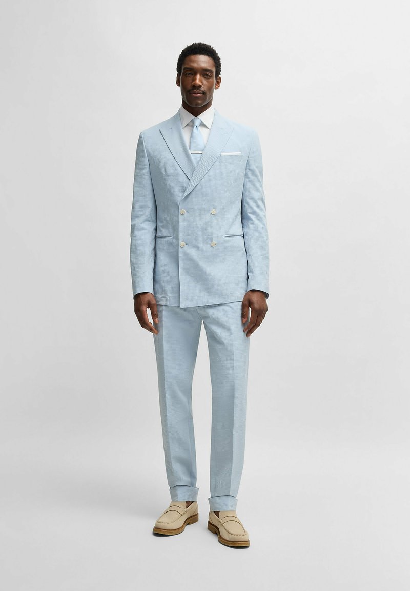 Ljusblå dubbelknäppt kostym med texturerad tyg, matchande slips, vit skjorta och beige loafers. Kostymen har två framfickor.