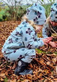 Fleeceoverall och matchande hatt i ljusblått med mönster av tvättbjörnar och träd, tillsammans med svarta vattentäta stövlar, omgiven av höstlöv.