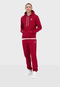 Felpa rossa con cappuccio, con lacci bianchi e tasca a marsupio, abbinata a pantaloni joggers rossi. Presenta un piccolo logo sul petto e sulla coscia.