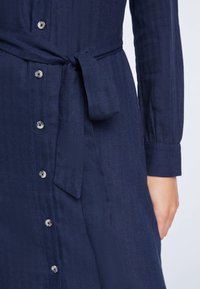 Granatowa sukienka z guzikami, uszyta z teksturowanej tkaniny, z paskiem w talii oraz długimi rękawami zakończonymi guzikami. Guziki są jasne.