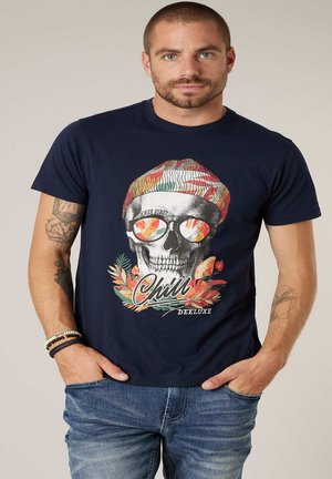 Homme portant un t-shirt bleu marine avec un dessin de crâne portant un chapeau coloré et des lunettes de soleil reflétant des feuilles tropicales, debout les mains dans les poches.