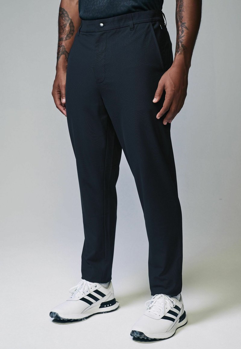 Schwarze, maßgeschneiderte Hose mit subtiler Textur, ausgestattet mit einem Vorderfalten, seitlichen Taschen und einem Knopfverschluss, getragen mit weißen Sportschuhen.