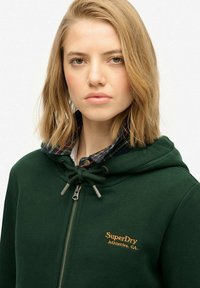 Mujer rubia vistiendo un hoodie de SuperDry verde oscuro sobre una camisa de cuadros, mirando al frente con una expresión neutral sobre un fondo sencillo.